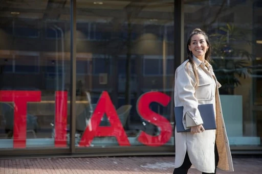 A woman in a light coat smiles while walking outside a building with large red letters on the window, holding a laptop.