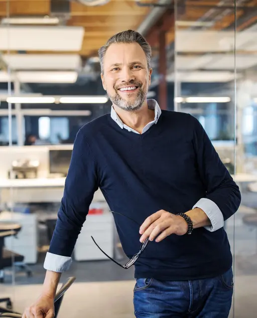 Smiling man with glasses in a modern office, leaning on a chair.