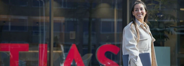 Smiling woman with a laptop walks by a building with large "TIAS" signage, suggesting a business or educational environment.