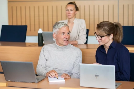 Three people in a classroom setting, two seated at desks with laptops having a discussion, and one sitting behind them, listening attentively.