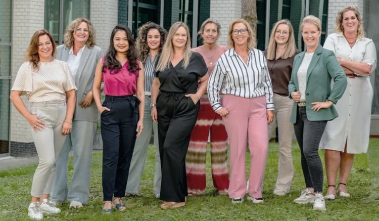 A diverse group of women smiling and standing together on a grassy area in front of a building.