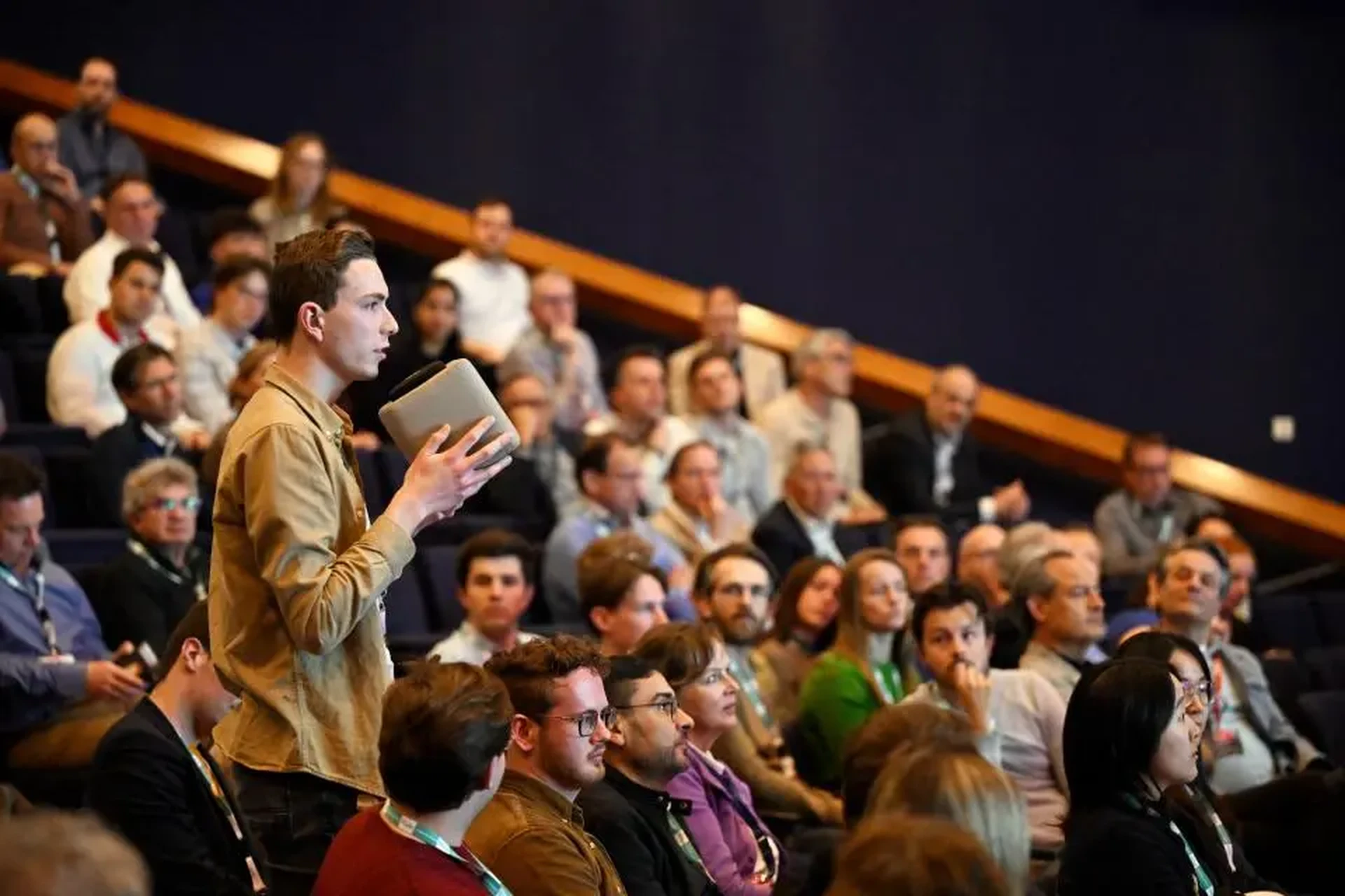 Audience member asking a question with a microphone in a crowded lecture hall during a conference.