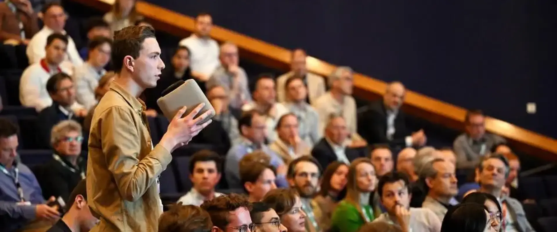 Audience member asking a question with a microphone in a crowded lecture hall during a conference.
