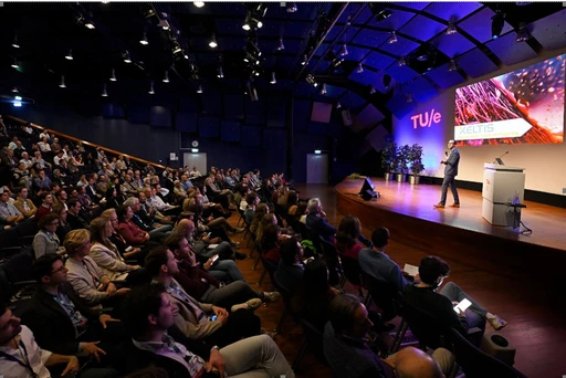 A speaker presents on stage to a large audience in a modern conference hall at TU/e.