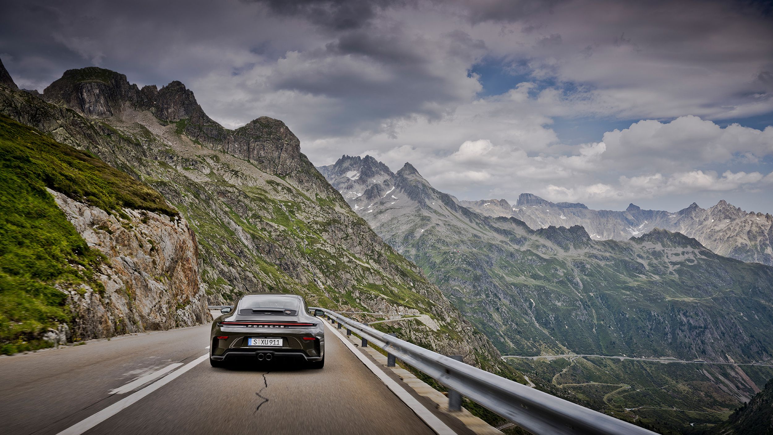 Porsche 911 GT3 Bergsport von hinten auf Alpenpassstrasse mit weitem Bergpanorama unter bewölktem Himmel.