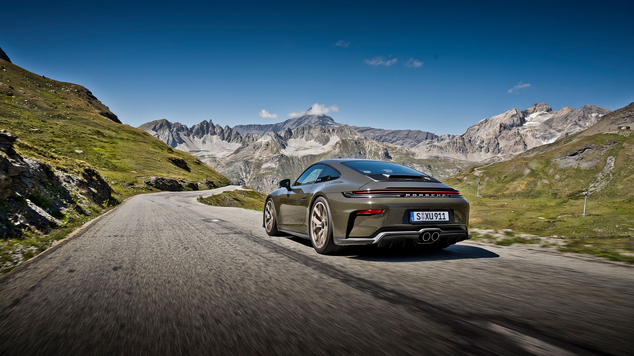 Porsche 911 GT3 Bergsport von hinten auf kurvenreicher Alpenpassstrasse mit grünen Wiesen und Bergpanorama.