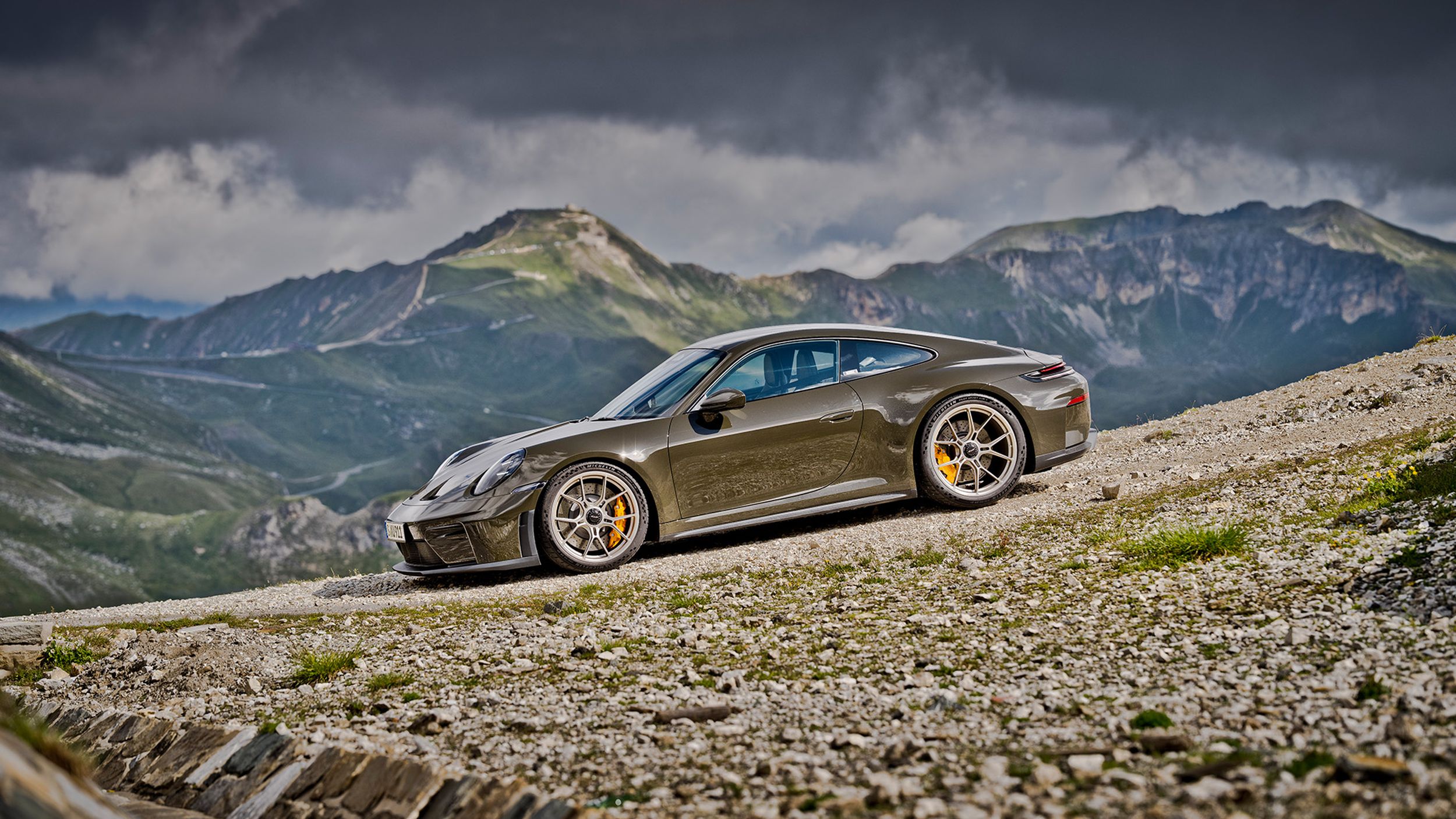 Porsche 911 GT3 Bergsport im Seitenprofil mit gelben Bremssätteln auf Schotterweg vor weitem Bergpanorama.