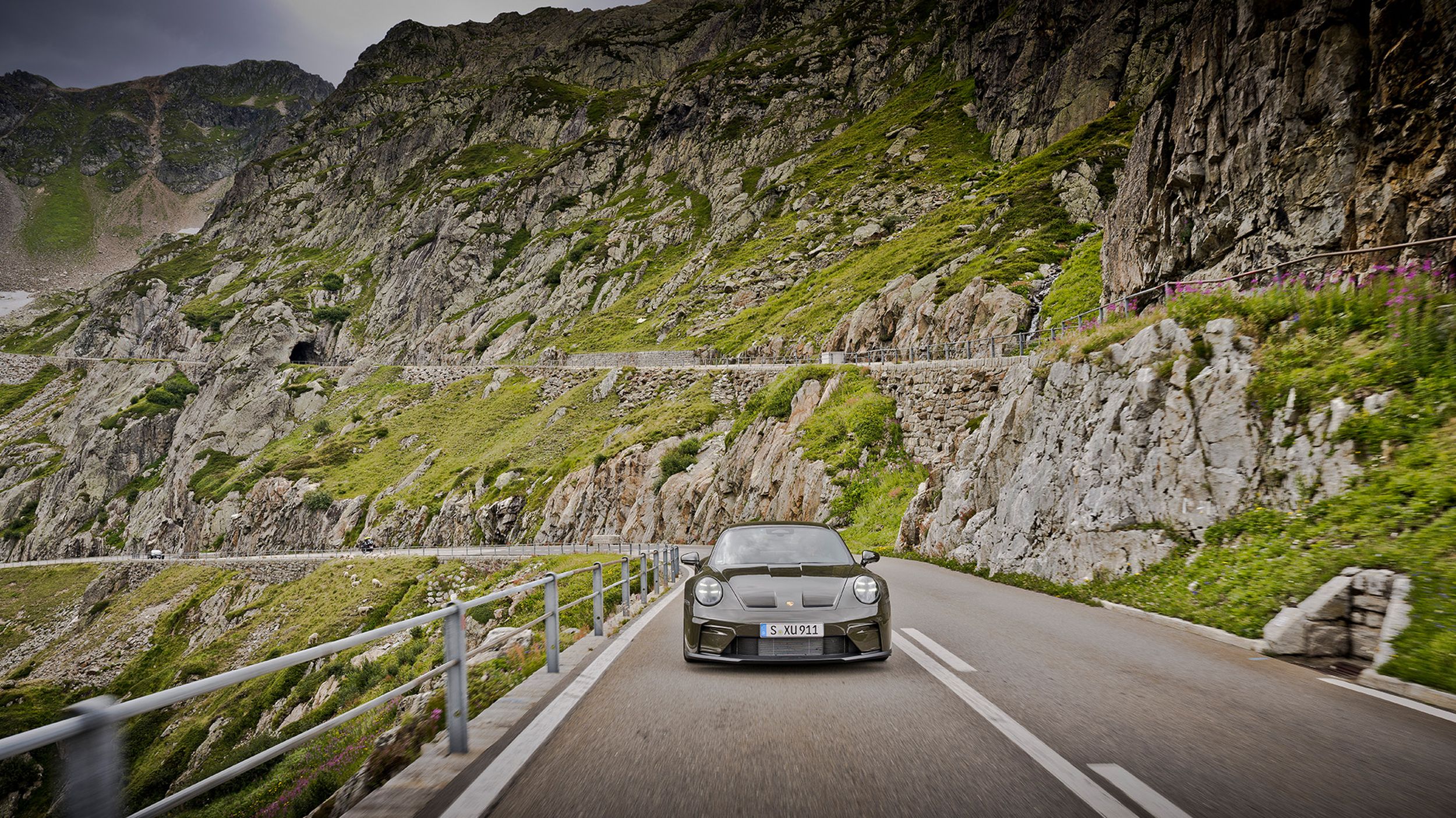 Porsche 911 GT3 Bergsport von vorne auf einer engen Alpenpassstrasse mit grünen Felshängen unter bewölktem Himmel.