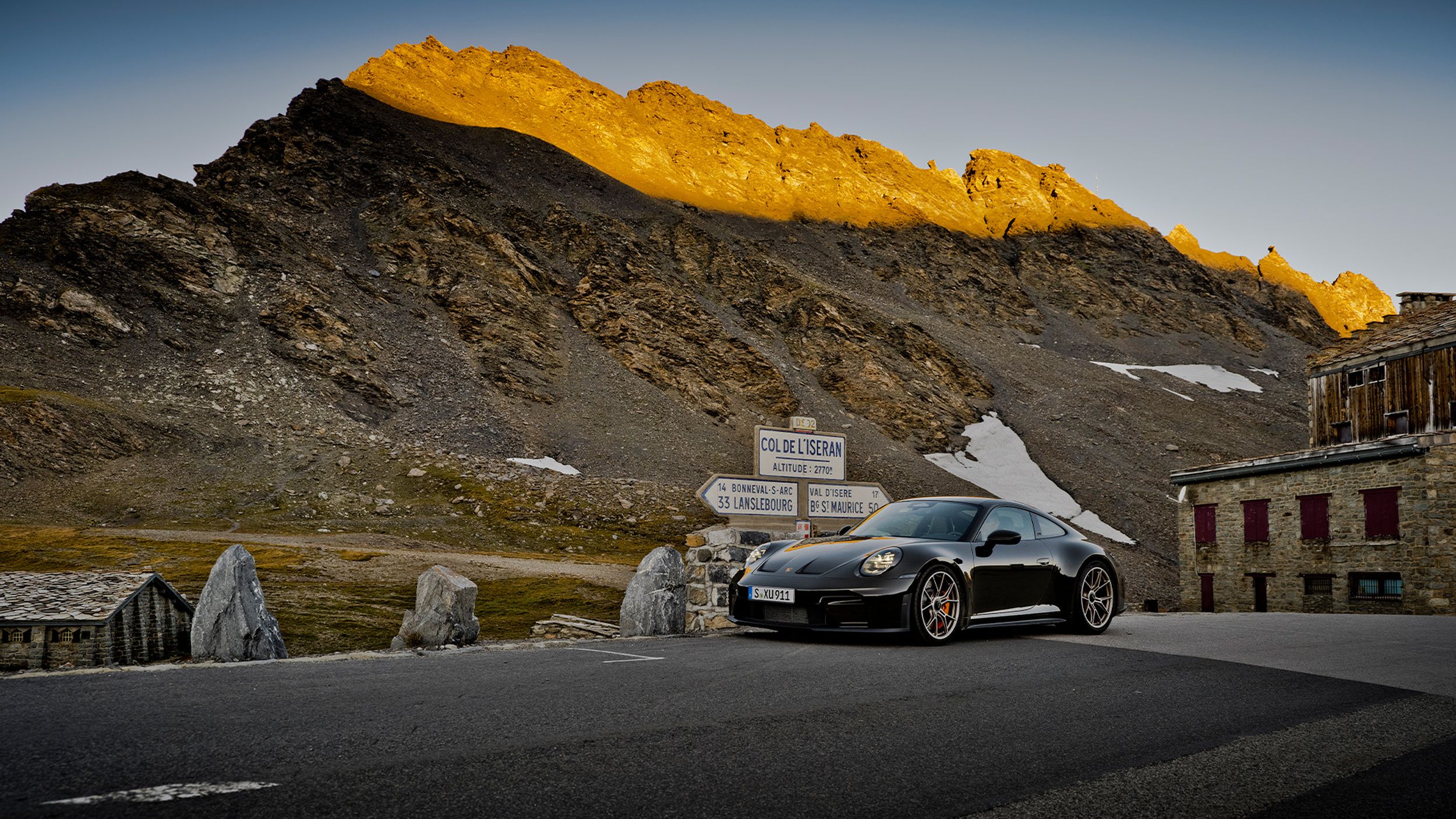 Porsche 911 GT3 Bergsport am Col de l'Iseran bei goldenem Sonnenuntergang über felsigem Alpengipfel.