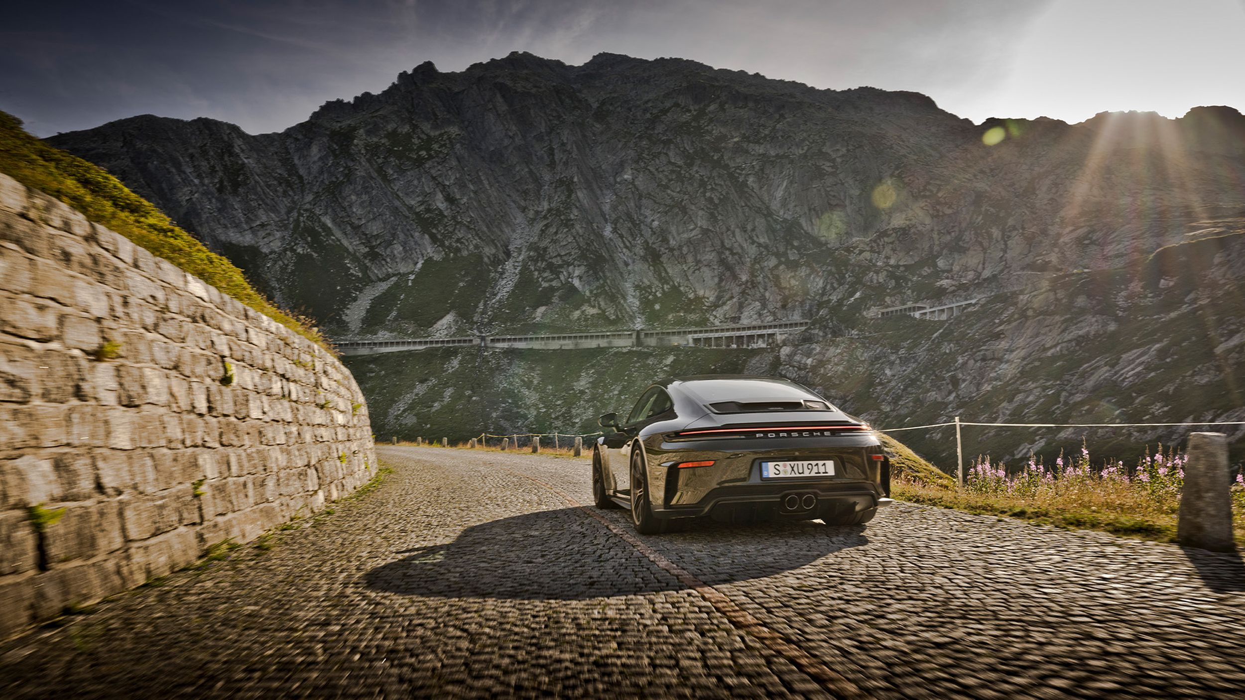 Porsche 911 GT3 Bergsport von hinten auf Kopfsteinpflaster neben Steinmauer im Gegenlicht vor felsigen Alpen.
