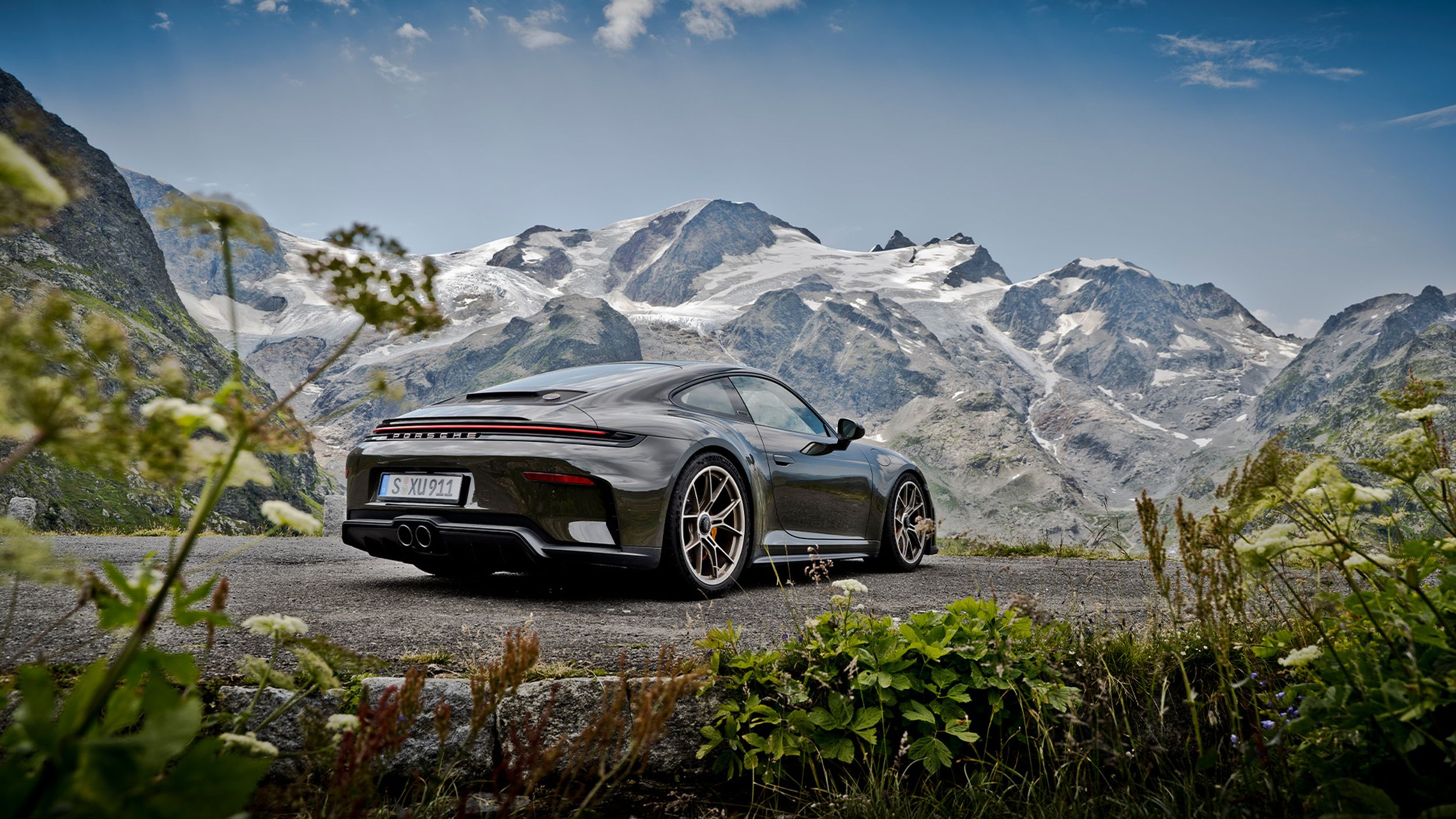 Porsche 911 GT3 Bergsport von hinten auf einer Bergstrasse mit Wildblumen und schneebedeckten Gipfeln.