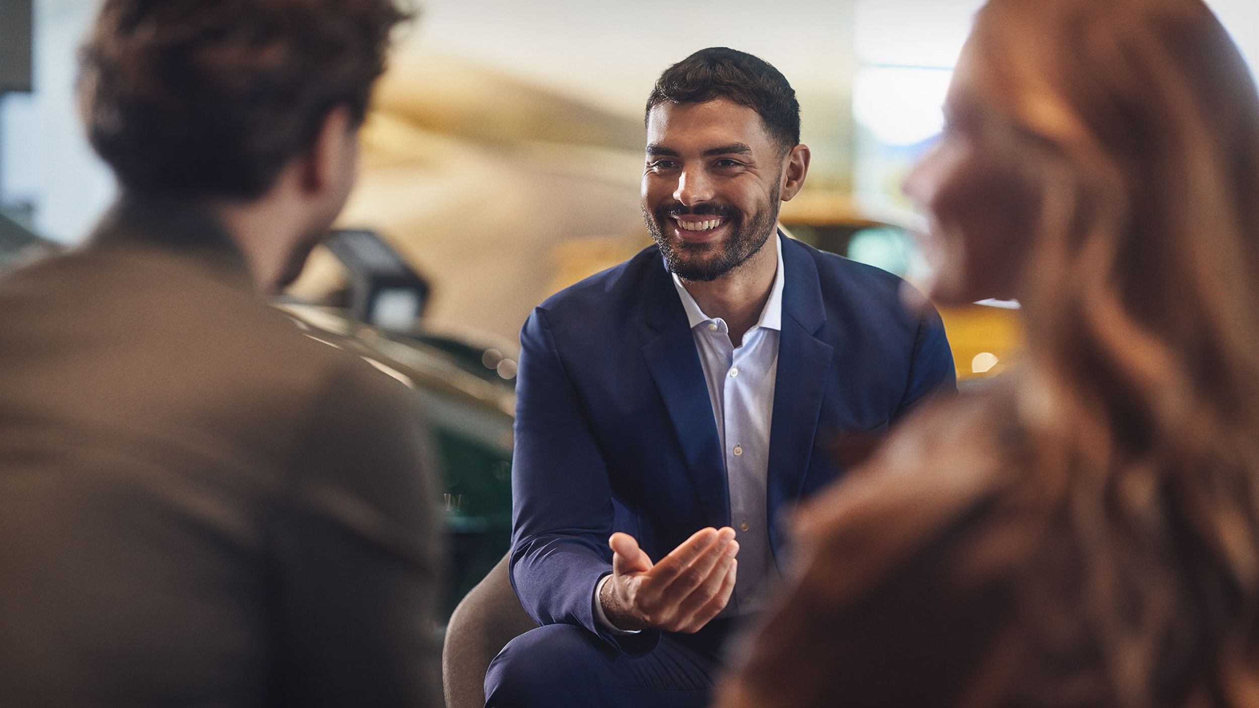 Porsche staff member facing two customers in a consultation at a Porsche Centre, with a green and yellow Porsche in the background.