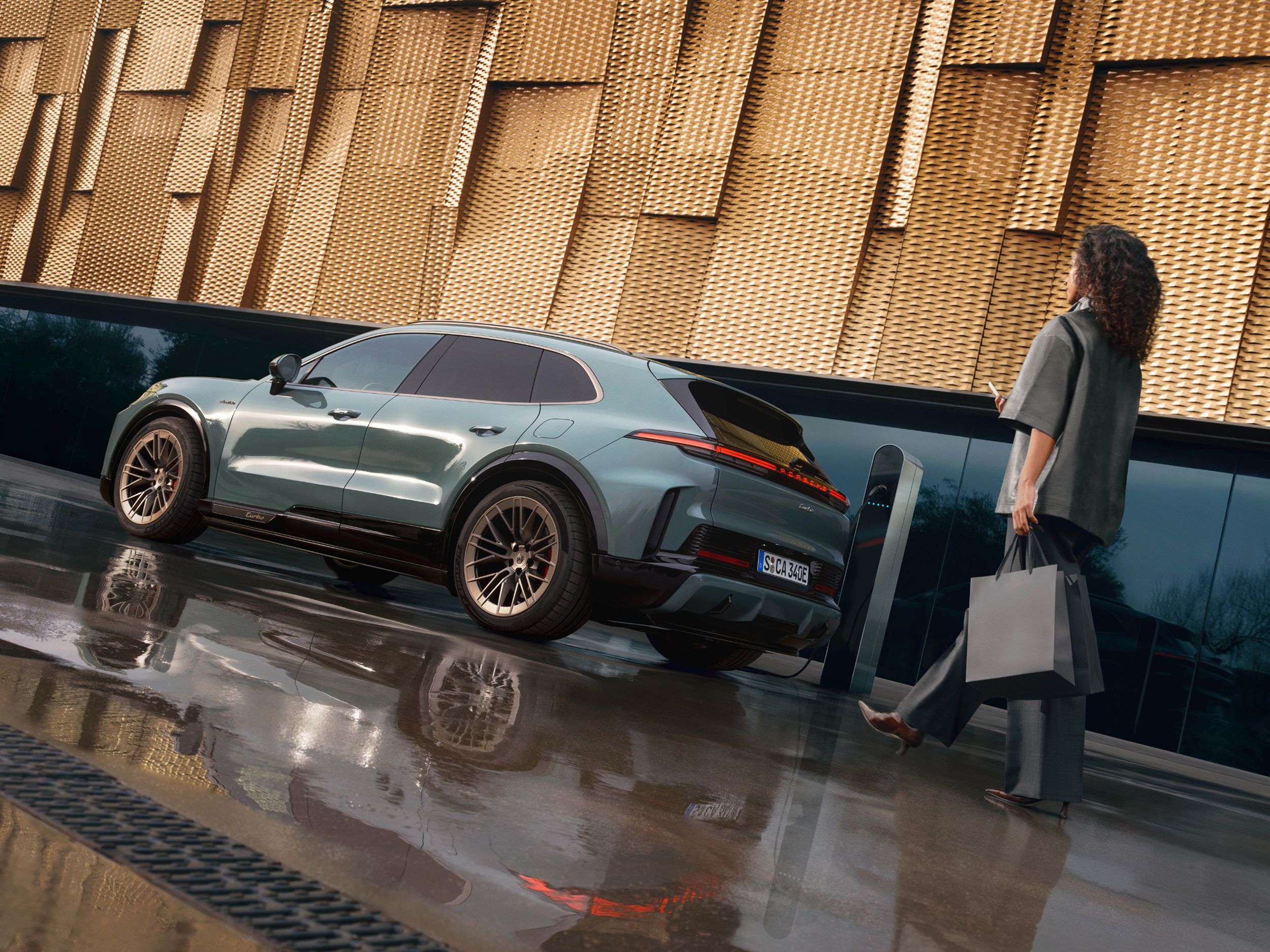 A Porsche Cayenne Turbo is charging at a charging point in front of a wall decorated with gold-coloured metal sheeting. The ground is wet. A woman carrying bags is walking towards the car.