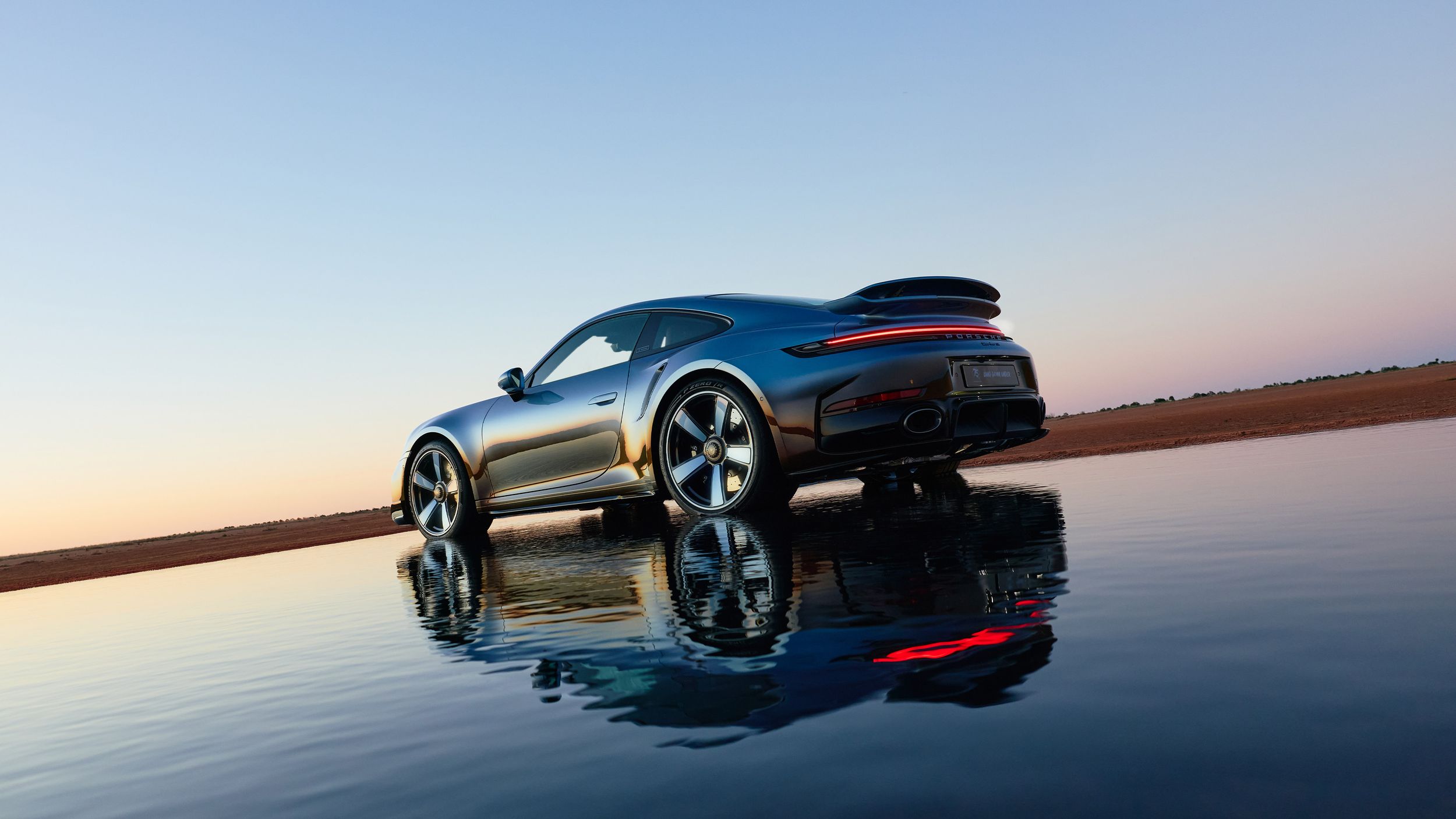 Side shot of the Porsche 911 Turbo S Land Down Under being reflected in a pool of water in a desert environment.