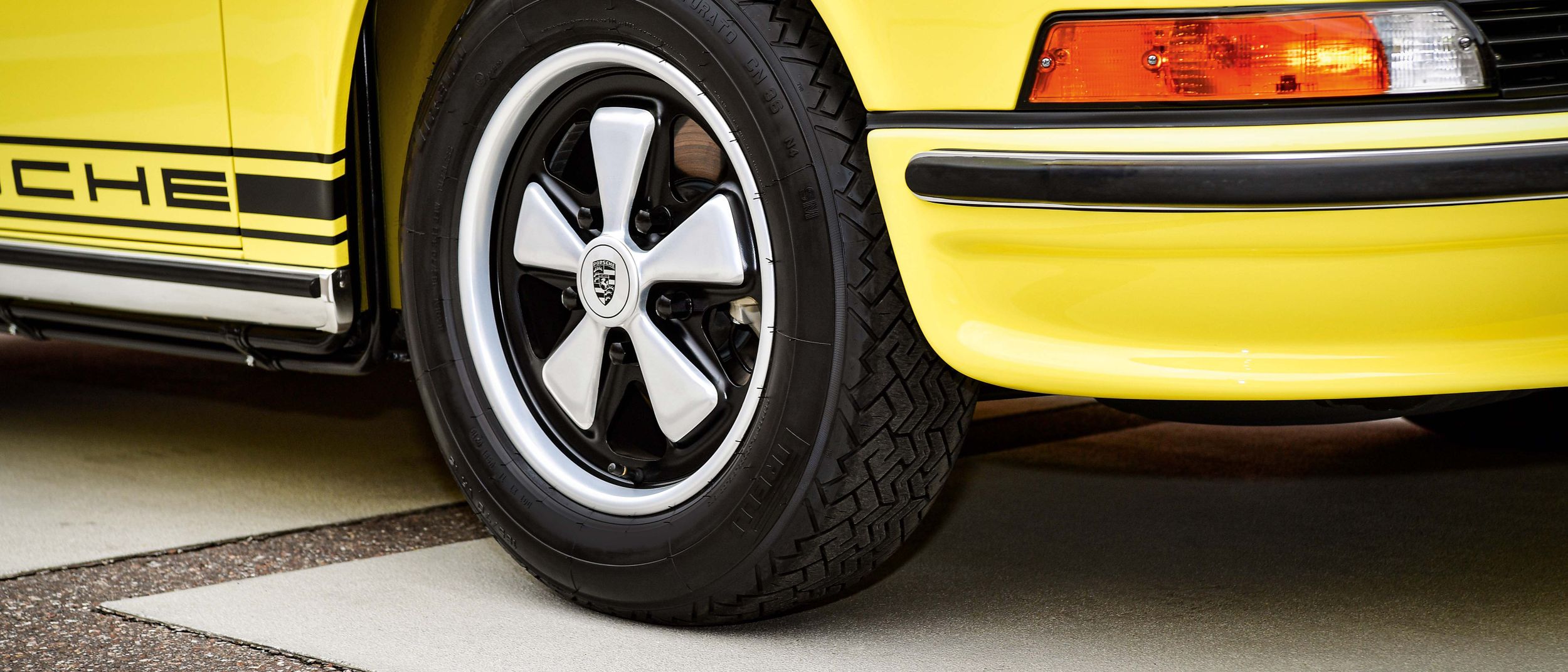 Close-up of the front section of a yellow classic car with black tyres, five-spoke rims, silver classic hubcaps and bumper.