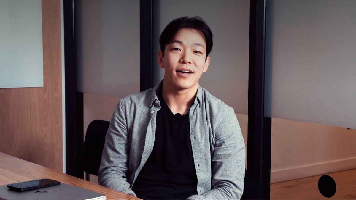 A person sitting at a desk, wearing a gray shirt over a black top, with a laptop nearby, looking towards the camera and smiling slightly.