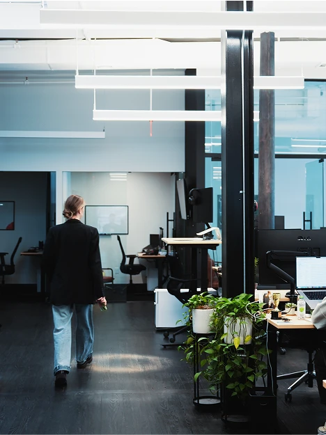 Open office space with modern decor, a person walking, desks with computers, and a plant on a stand. Bright lighting and large windows.