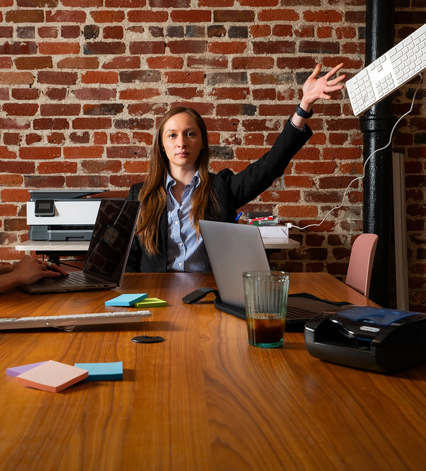 A woman in an office setting dramatically gestures with a keyboard in hand, surrounded by laptops, a printer, and colorful sticky notes.