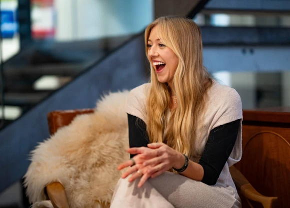Smiling woman with long blonde hair sits on a chair with a fur throw, clapping hands, in a modern, warmly lit interior.