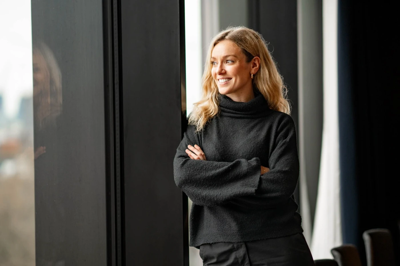 Woman with blonde hair in a black sweater, smiling and standing by a window with arms crossed, looking outside.