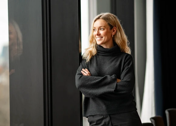 Woman with blonde hair in a black sweater, smiling and standing by a window with arms crossed, looking outside.