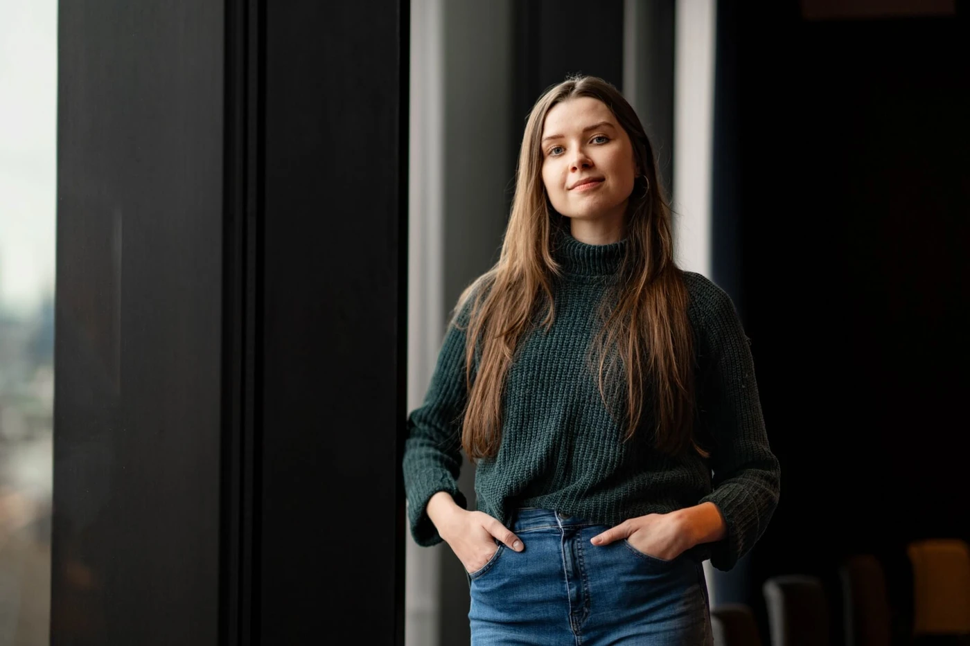 Woman with long hair in a dark sweater and jeans stands by a large window, hands in pockets, looking confidently at the camera.