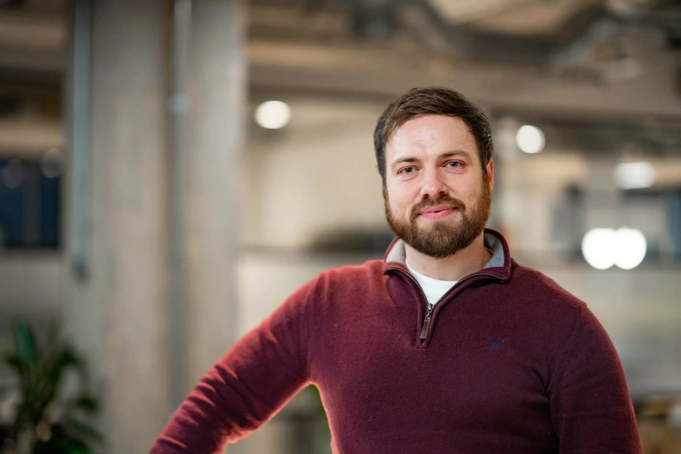 Man with a beard in a maroon sweater stands in a modern office setting, smiling at the camera.