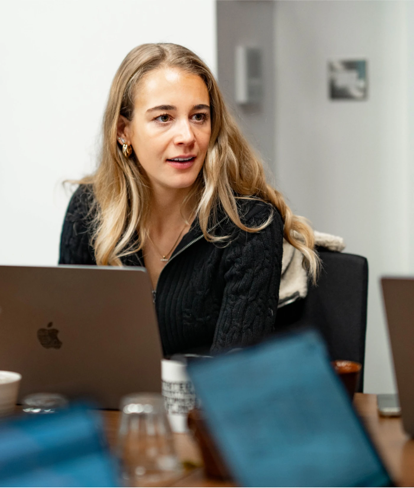 Woman with long blonde hair sits at a table with a laptop, engaged in conversation, in a modern office setting.