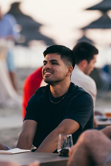 A person sitting outdoors at a beachside setting, smiling and looking away. Blurred background with people and umbrellas.