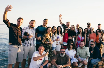 Group of people posing cheerfully by the water, some holding drinks, with a clear sky in the background.