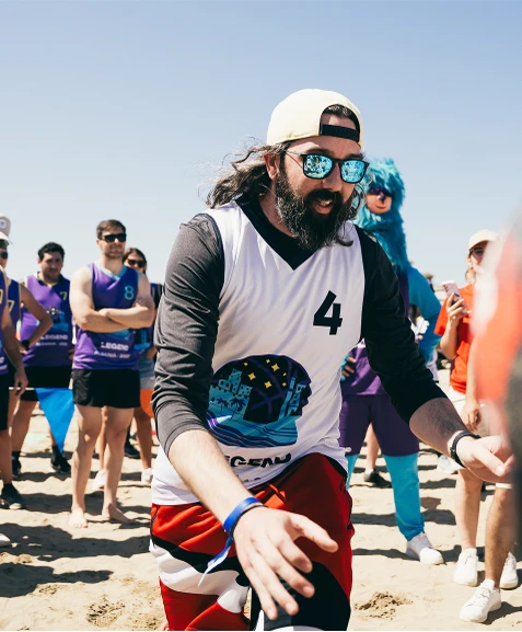 Man with a beard and sunglasses playing beach sports, wearing a jersey with the number 4. People and a blue mascot are in the background.