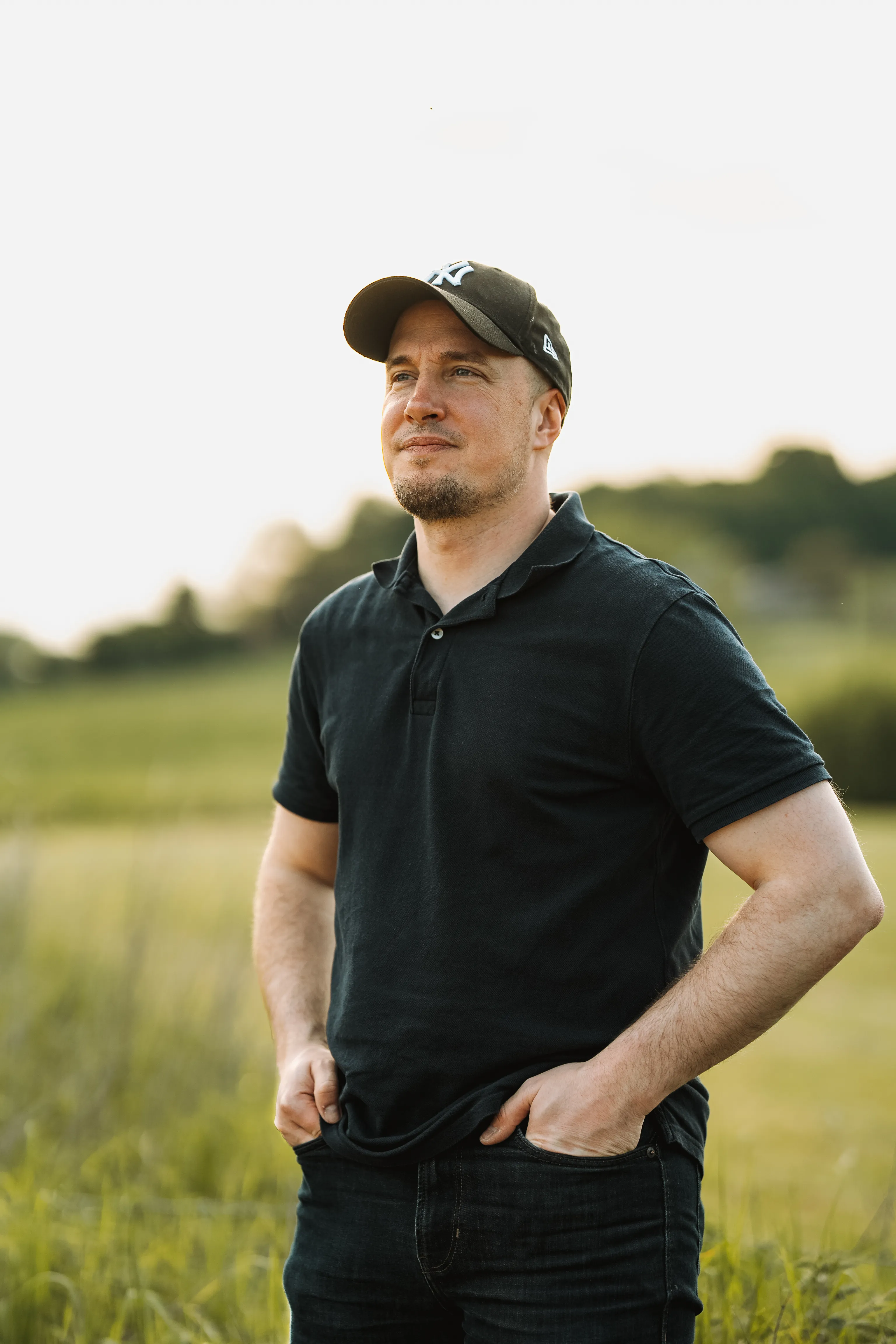 un homme en t-shirt noir et avec une casquette et vu de face avec les mains en poche