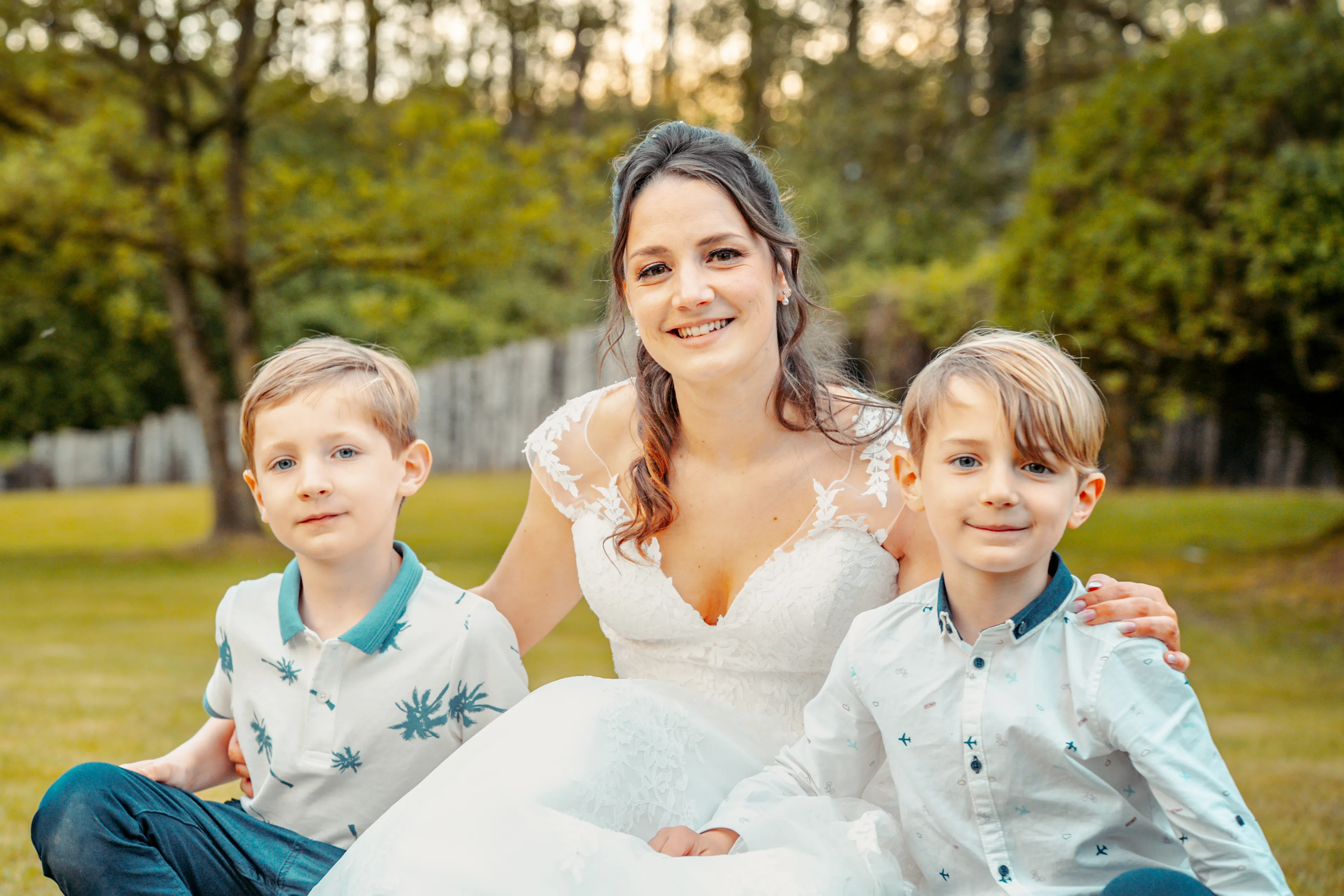 une mariée est assise avec deux garçons dans l'herbe, elle a ses mains sur leurs épaules