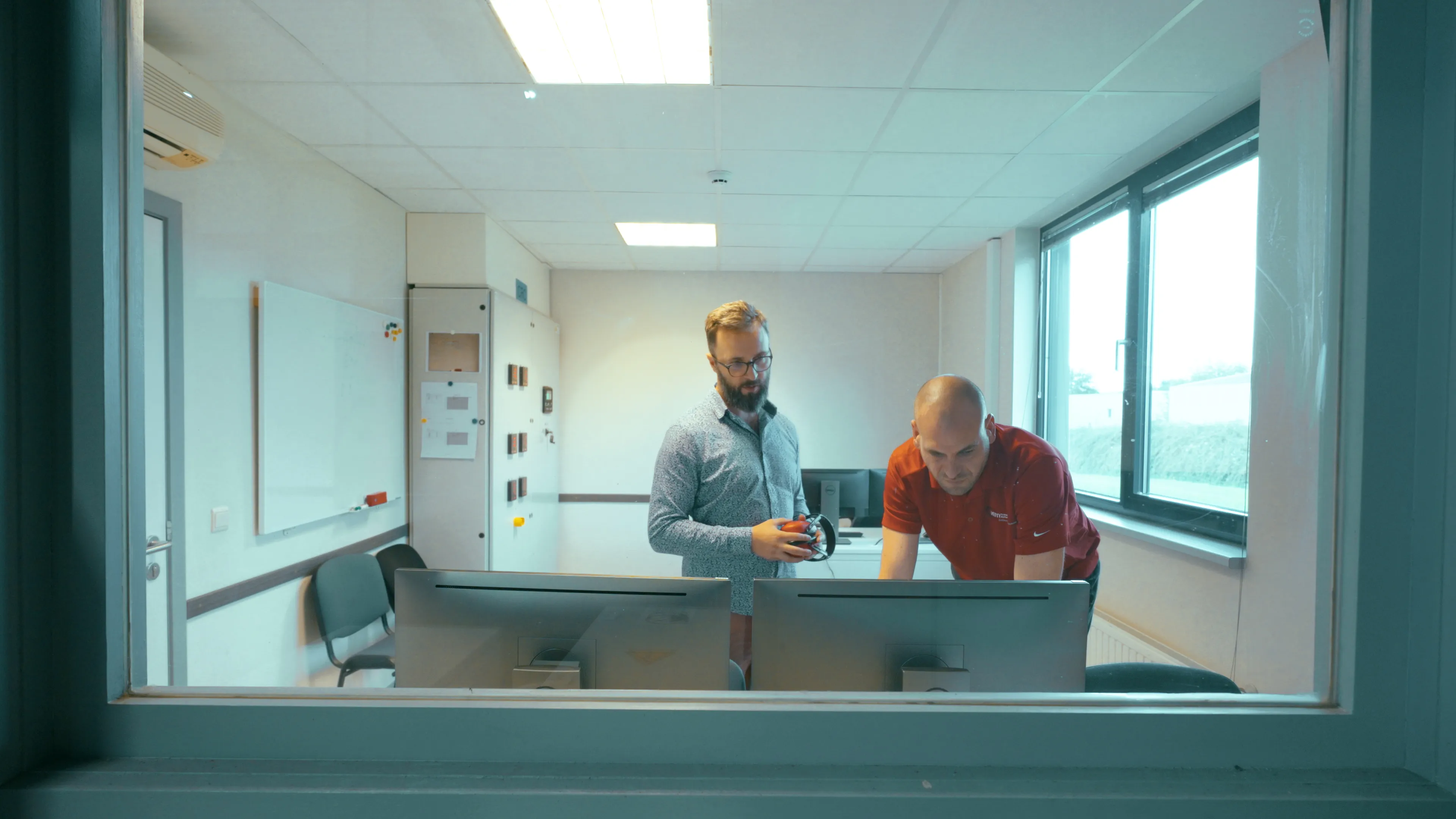 deux hommes sont visibles derrière une vitre, ils regardent un écran d'ordinateur