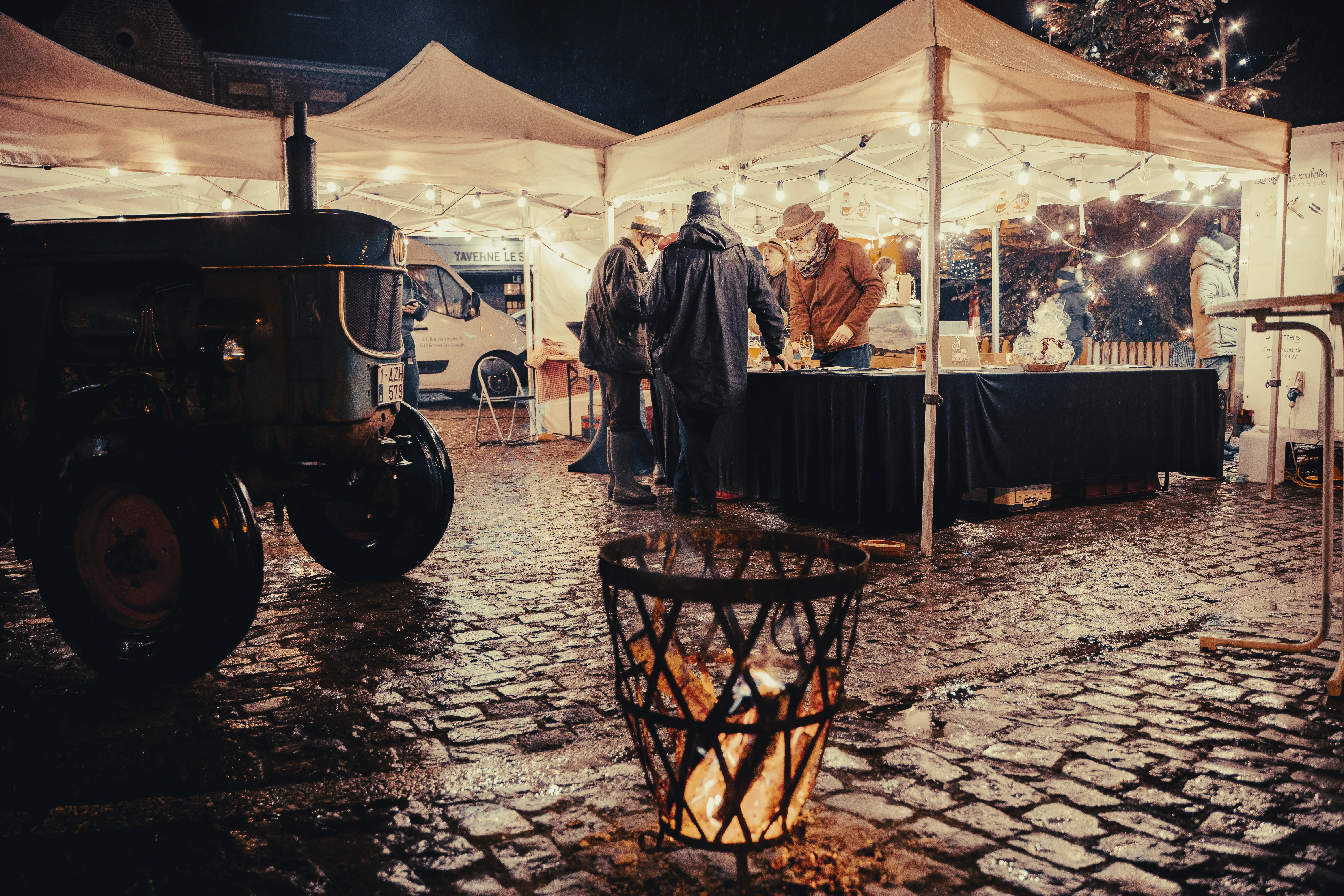 en soirée, un groupe de personnes est en-dessous d'une tente illuminée, ils sont debout autour d'une table - un braséro allumé est au premier plan