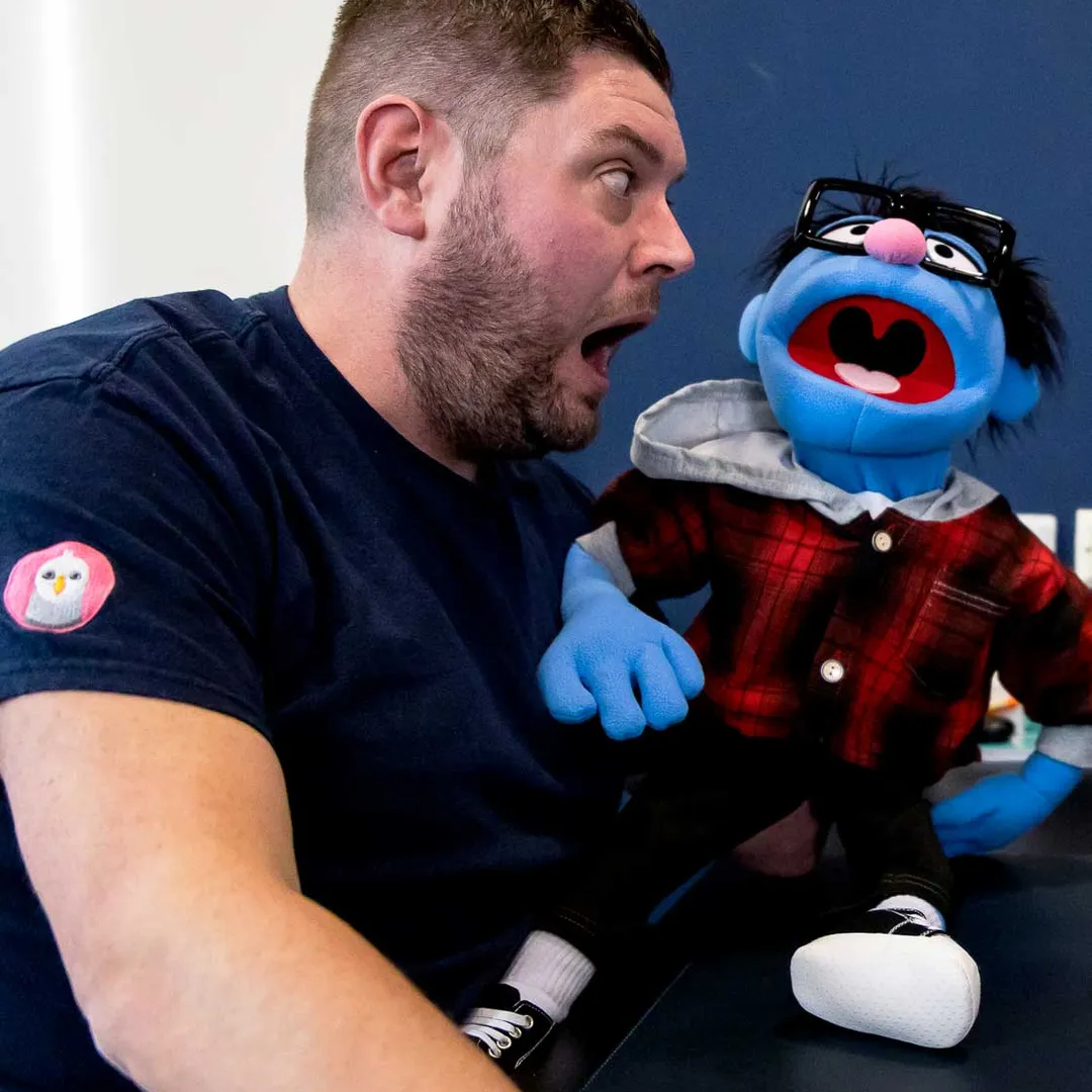 A man playfully interacts with a blue puppet wearing glasses and a red plaid shirt, both with mouths open in surprise.