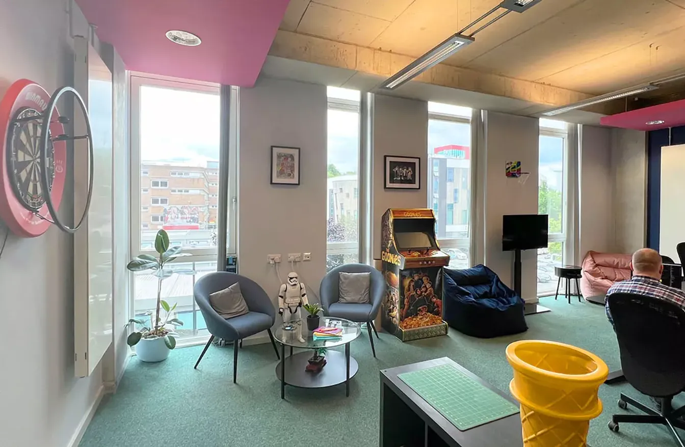 A modern lounge with large windows, arcade machine, dartboard, small seating area, and decor. A person sits in the corner.