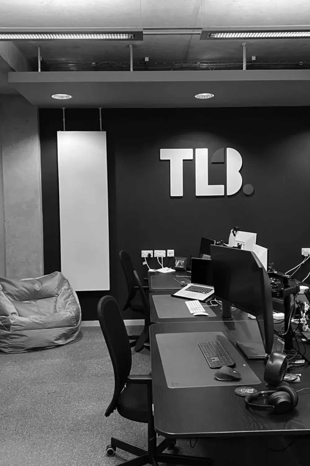 Black and white image of a modern office with desks, computers, a bean bag, arcade machine, and "TLB" logo on the wall.