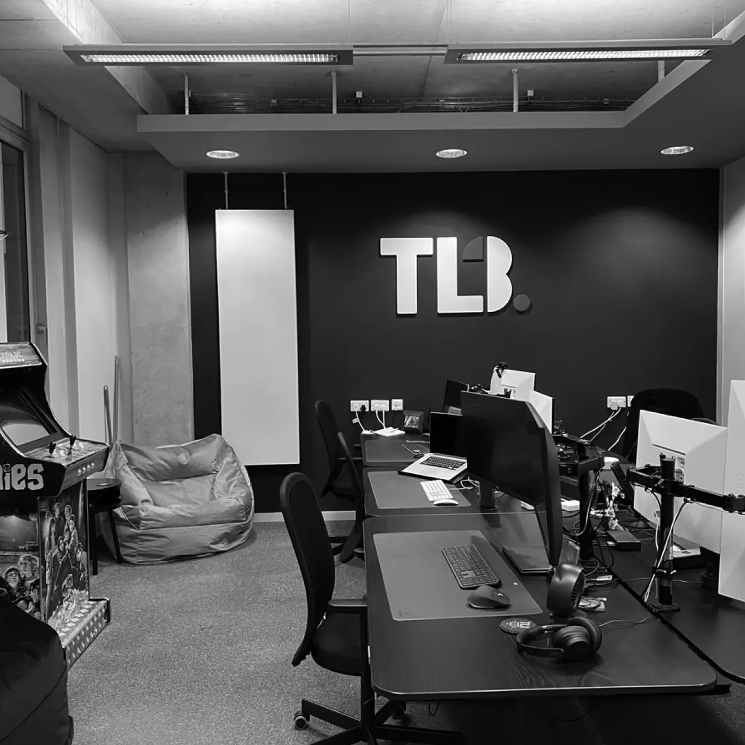 Black and white image of a modern office with desks, computers, a bean bag, arcade machine, and "TLB" logo on the wall.