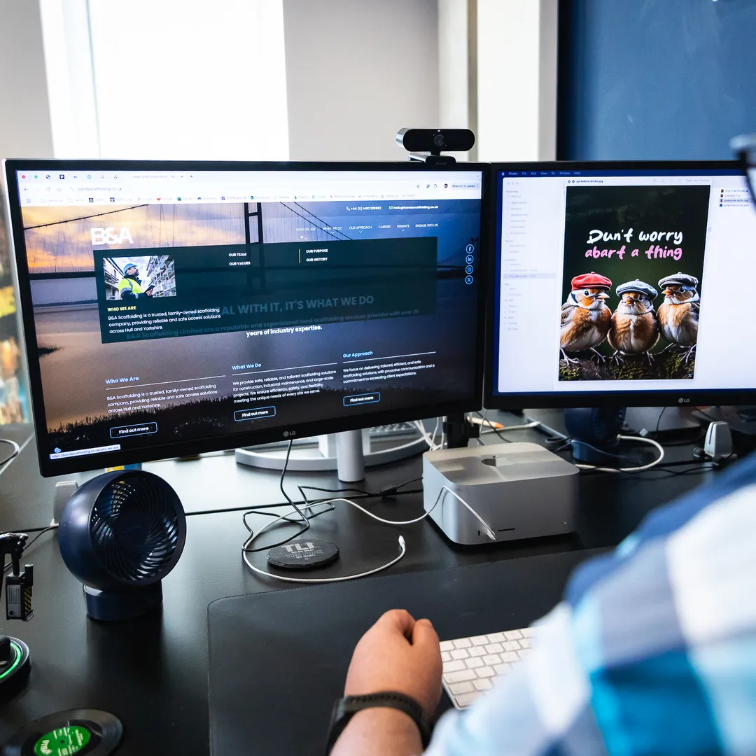 A person in a plaid shirt works at a dual-monitor setup displaying a website and an image of meerkats with the text "Don't worry about a thing."
