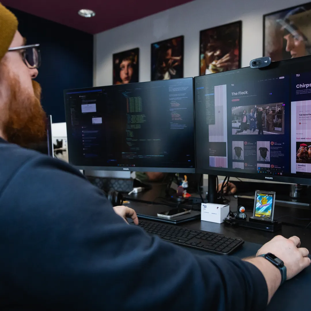 Person wearing a beanie working on dual monitors, with code and graphics displayed, in a modern office with posters on the wall.