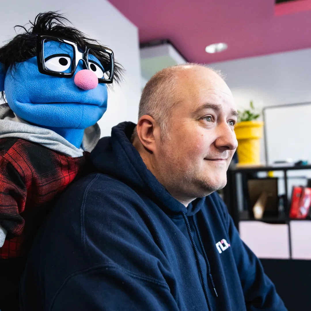 A man in a hoodie sits next to a blue puppet with glasses and a plaid shirt in an office setting.
