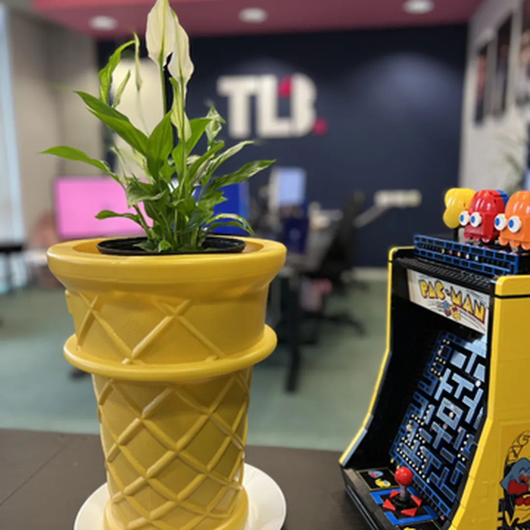 A peace lily in a yellow, ice cream cone-shaped pot beside a mini Pac-Man arcade machine on a table.