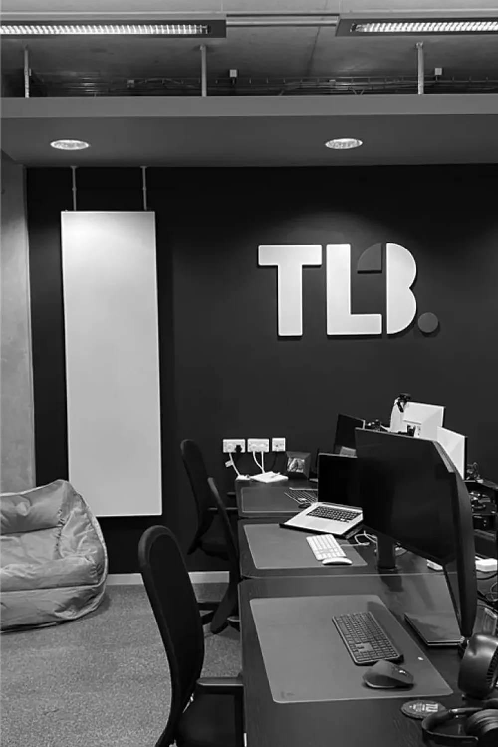 Black and white image of a modern office with desks, computers, a beanbag, and "TLB" logo on the wall.
