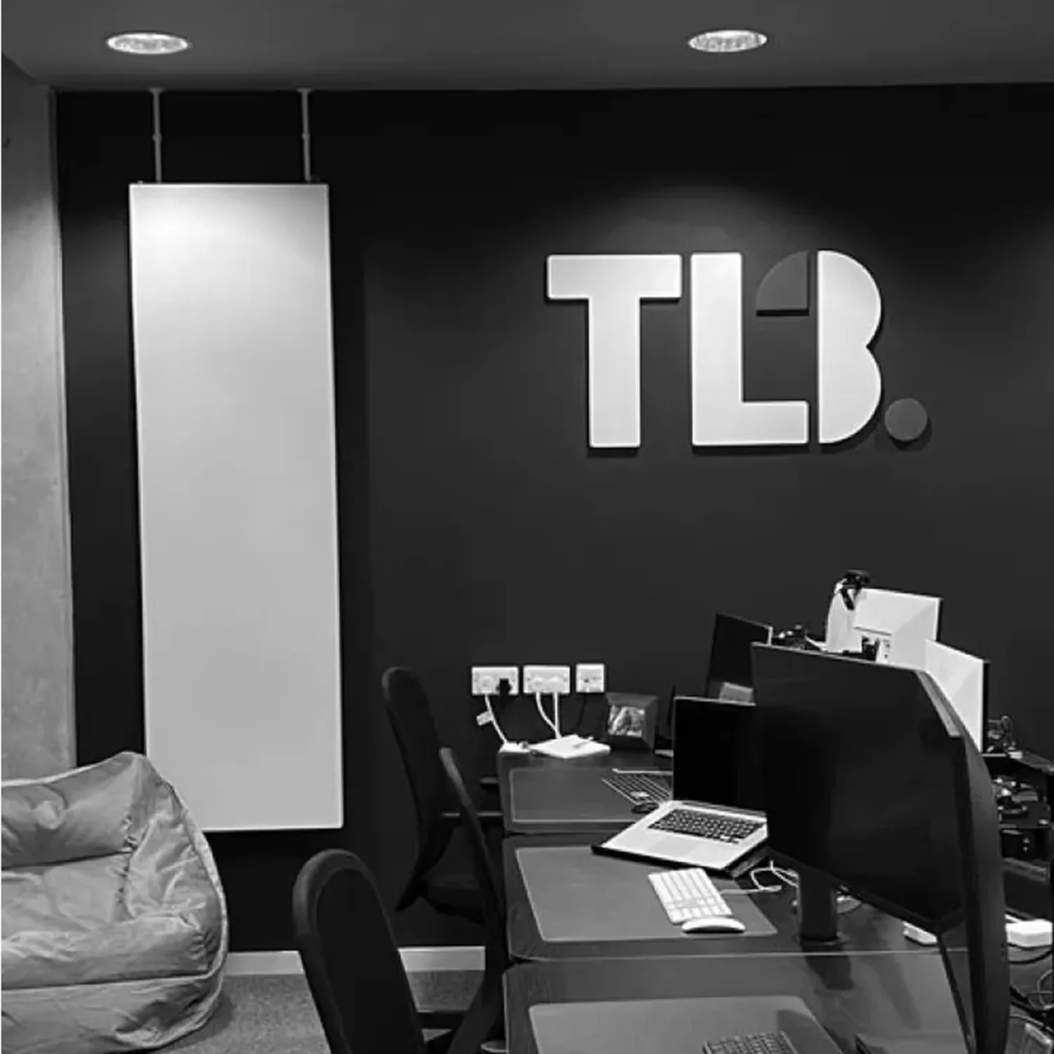 Black and white image of a modern office with desks, computers, a beanbag, and "TLB" logo on the wall.