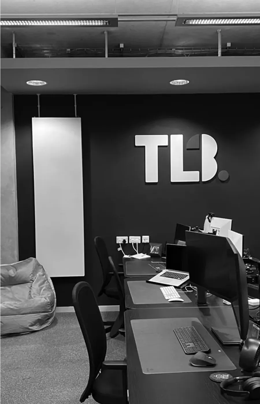 Black and white image of a modern office with desks, computers, a beanbag, and "TLB" logo on the wall.