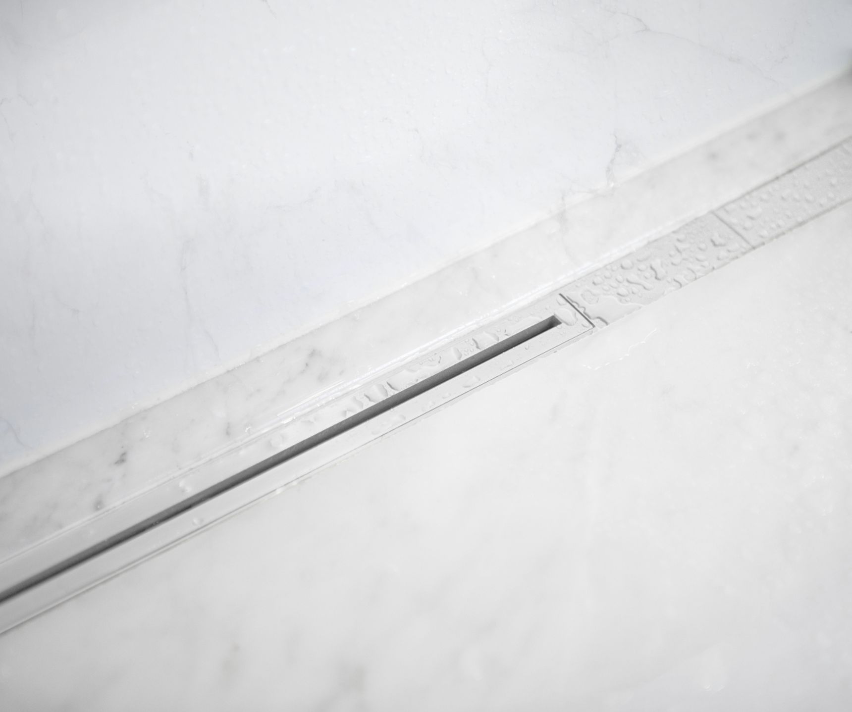 Close-up of a sleek, modern linear shower drain set in a marble-tiled floor, with water droplets visible on the surface.
