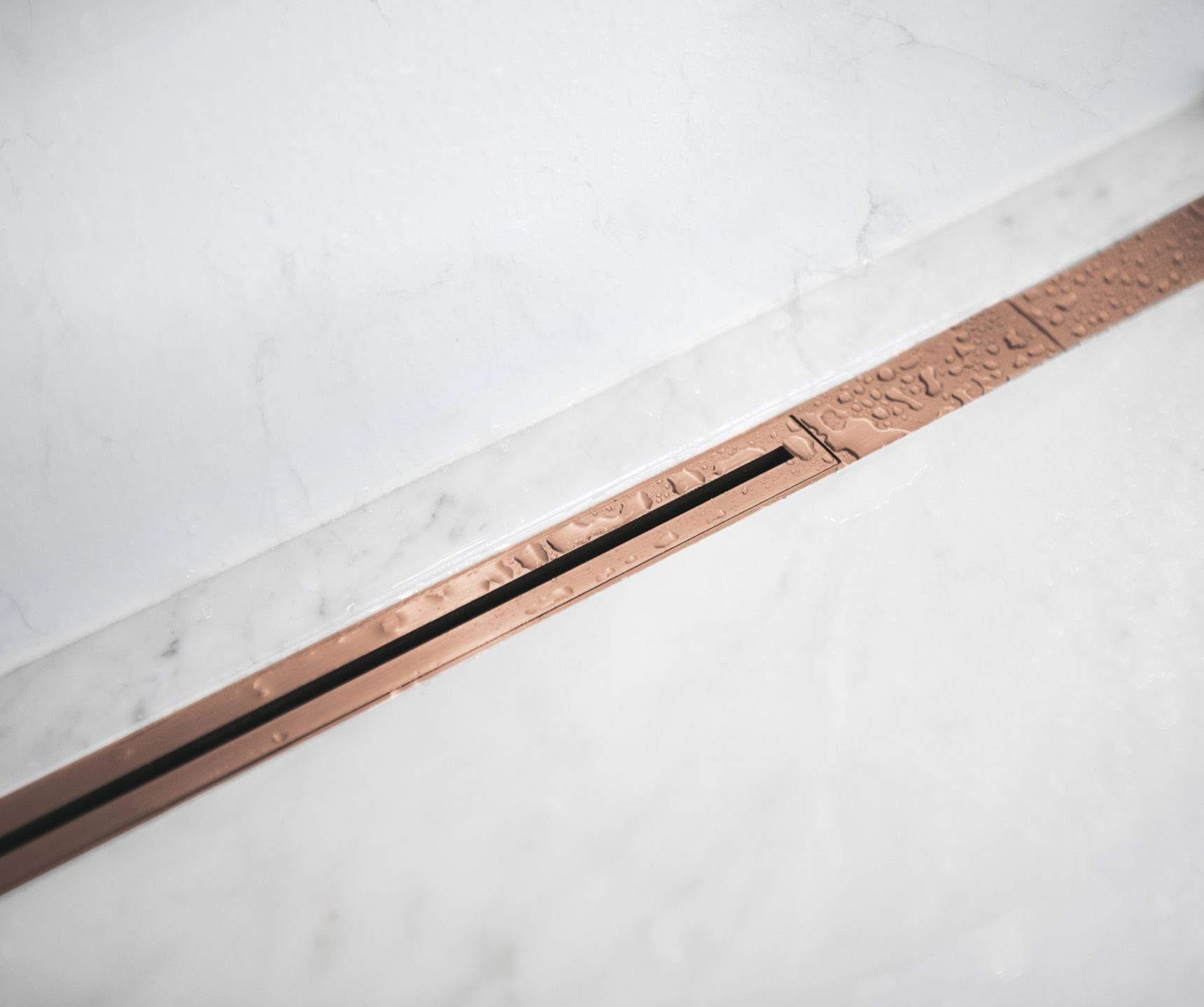 Close-up of a sleek, modern shower drain with a copper finish embedded in a white marble floor, featuring water droplets.