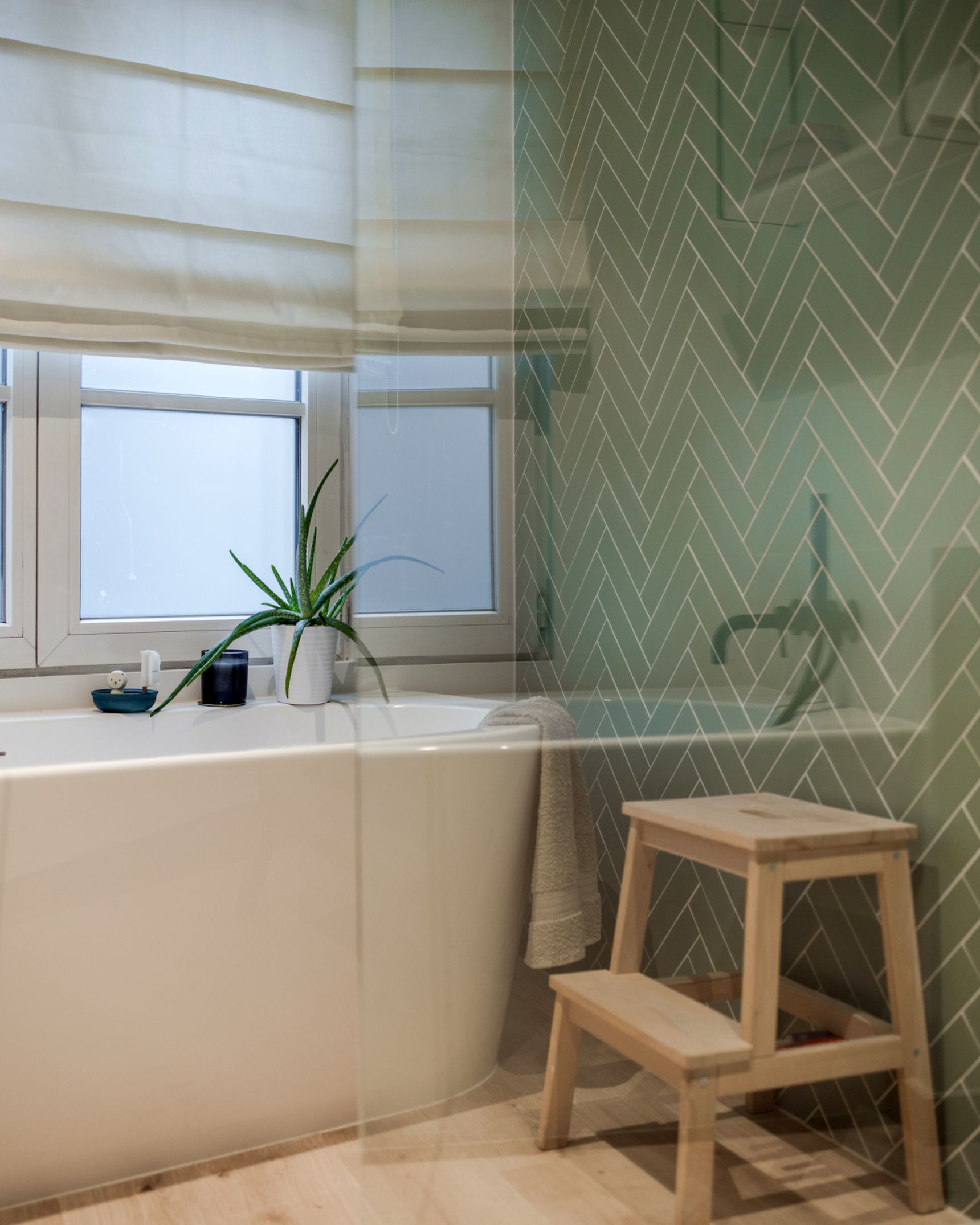 Modern bathroom with a white bathtub, wooden step stool, green herringbone tile wall, and a potted plant near a frosted window.