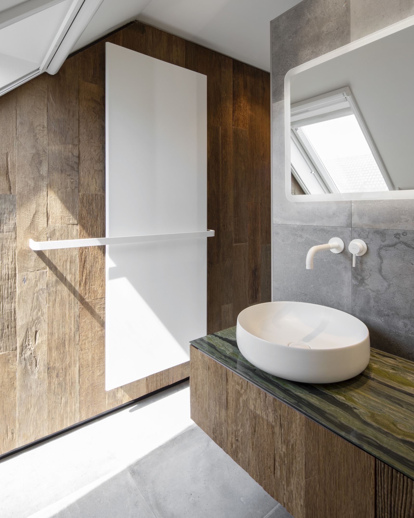 Modern bathroom with a round white sink on a wooden vanity, angled mirror, and skylight. Wood-paneled wall and gray stone flooring.